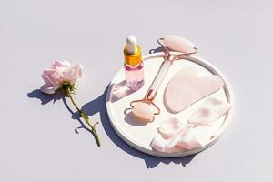 Rose pétrole cosmétique bouteille, gua sha grattoir, quartz rouleau masseur sur céramique plateau avec Rose pétales. Accueil se soucier, massage. photo