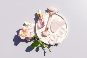 bouteille de cosmétique Rose huile, quartz masseur, gua sha grattoir sur blanc céramique plateau pour Accueil massage et soins auto-administrés. Haut voir. photo