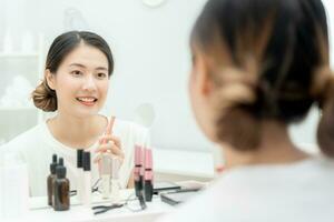 magnifique asiatique femme asseoir dans de face de une miroir et sourire sur se maquiller. visage de une en bonne santé femme appliquant se maquiller. publicité, mode de vie , produits de beauté, maquillage accessoires, beauté activité, esthéticien photo