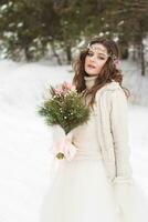 magnifique la mariée dans une blanc robe avec une bouquet dans une couvert de neige hiver forêt. portrait de le la mariée dans la nature photo