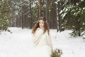 magnifique la mariée dans une blanc robe avec une bouquet dans une couvert de neige hiver forêt. portrait de le la mariée dans la nature photo