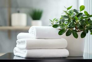 une empiler de duveteux blanc les serviettes est assis soigneusement sur une en bois table dans une salle de bains. suivant à le les serviettes est une petit, mis en pot plante avec vert feuilles. le salle de bains est nettoyer et rangé photo