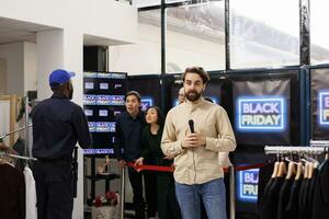 Jeune homme la télé journaliste couvrant nouvelles temps réel, vivre diffusion pendant noir Vendredi ventes. correspondant permanent à boutique entrée parlant dans microphone fabrication vivre nouvelles à propos achats la démence photo