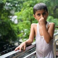 peu enfant en jouant dans été pluie dans maison balcon, Indien intelligent garçon en jouant avec pluie gouttes pendant mousson pluvieux saison, enfant en jouant dans pluie photo