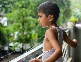 peu enfant en jouant dans été pluie dans maison balcon, Indien intelligent garçon en jouant avec pluie gouttes pendant mousson pluvieux saison, enfant en jouant dans pluie photo