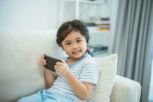 asiatique enfant fille à la recherche en utilisant et toucher mobile téléphone écran sur canapé canapé. bébé souriant marrant temps à utilisation mobile téléphone. aussi beaucoup écran temps. mignonne fille en train de regarder vidéos tandis que la télé, l'Internet dépendance. photo