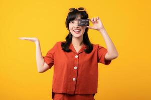 portrait Jeune magnifique asiatique femme content sourire habillé dans Orange vêtements montrant crédit carte isolé sur Jaune Contexte. Payer et achat achats Paiement concept. photo