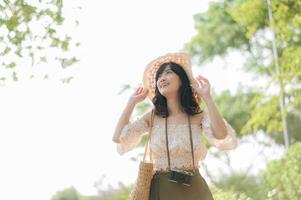 portrait de asiatique Jeune femme voyageur avec tissage chapeau et panier et une caméra sur vert Publique parc la nature Contexte. périple voyage mode de vie, monde Voyage explorateur ou Asie été tourisme concept. photo