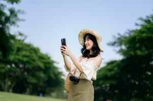 portrait de asiatique Jeune femme voyageur avec tissage chapeau, panier, mobile téléphone et caméra sur vert Publique parc Contexte. périple voyage mode de vie, monde Voyage explorateur ou Asie été tourisme concept. photo