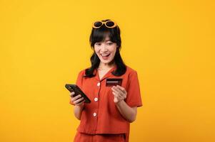 portrait Jeune magnifique asiatique femme content sourire habillé dans Orange vêtements en utilisant crédit carte achat achats en ligne sur téléphone intelligent isolé sur Jaune Contexte. Payer et achat Paiement concept. photo