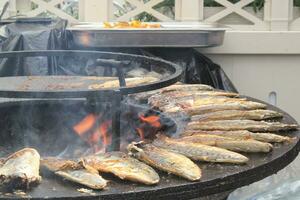 grillé poisson sur une en forme de bol plat gril avec Feu trou dans le centre. Extérieur Fruit de mer barbecue cuisson. pique-nique sur arrière-cour sur ouvert air. rue nourriture concept. en train de préparer poisson plus de ouvert feu de camp photo