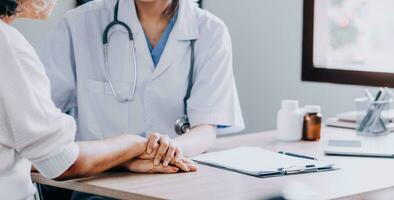 médecin donnant espoir. proche en haut coup de Jeune femelle médecin penché vers l'avant à souriant personnes âgées Dame patient en portant sa main dans palmiers. femme le concierge dans blanc manteau justificatif encourageant vieux la personne photo