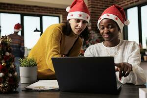 souriant collègues de travail dans Père Noël Chapeaux vérification annuel rapport résultats sur portable dans décoré de fête bureau. africain américain et caucasien collègues gérant projet à Noël veille photo