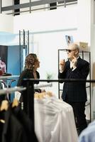 Sénior homme en essayant élégant des lunettes de soleil dans Vêtements magasin, nécessaire Aidez-moi de salle d'exposition employé. personnes âgées client à la recherche à Nouveau mode collection, achat à la mode vêtements dans moderne boutique photo