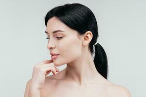 fermer beauté portrait de Jeune femme avec lisse en bonne santé peau, elle doucement touche sa visage avec sa les doigts sur lumière gris Contexte et sourit. soin de la peau concept photo