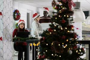 excité entreprise directeur profiter Noël esprit humeur, apportant paquet à travail plein de de fête ornements aidé par employé. affaires équipe chef orner lieu de travail avec Noël décorations photo