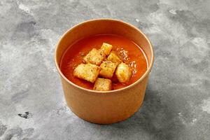 du froid Gaspacho soupe avec toasts, séché herbes dans papier bol sur gris pierre surface photo