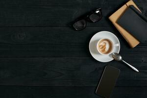 au dessus vue de intelligent téléphone avec carnet et tasse de latté café sur noir en bois Contexte. photo