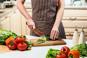 Jeune femme cuisine dans le cuisine à maison. une femme coupes une concombre et des légumes avec une couteau. photo