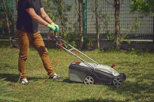 Masculin dans décontractée tenue et gants est Coupe vert herbe avec moderne pelouse tondeuse sur le sien cour. jardinage se soucier équipement et prestations de service. ensoleillé journée photo