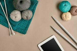 gris et turquoise tricot laine, tricot aiguilles et une tablette avec une noir écran sur beige Contexte. Haut voir. copie espace photo