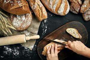 femme mains Coupe une pain de pain sur rustique en bois conseil, avec blé oreilles et couteau, Haut voir. photo