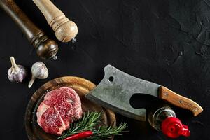 pièce de du boeuf filet, avec hache pour Coupe et couper Viande, épices étaient cuisine - Romarin, poivre, sel, Ail. sur vieux en bois planche sur pierre noir tableau. photo