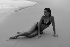 élégant femme avec parfait figure posant sur tropical plage. mensonge sur sable. portant élégant noir maillots de bain. noir et blanche. photo