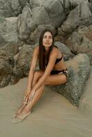 séduisant brunette femme avec parfait figure posant sur tropical rocheux plage. portant élégant noir maillots de bain. photo