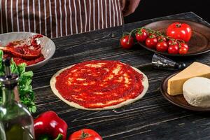 une Masculin main diffusion tomate purée sur une Pizza base avec cuillère sur un vieux en bois Contexte photo