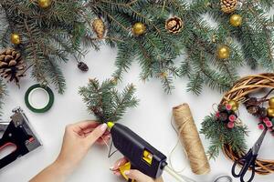 Haut vue de femelle mains faire une Noël couronne. emballé cadeaux et volutes, épicéa branches et outils sur blanc tableau. photo