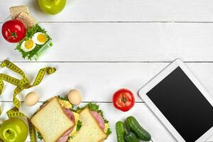 blanc tablette ordinateur avec une Vide écran sur le cuisine table avec sandwichs, Frais légumes, bouteille de l'eau et Pomme. le déjeuner photo