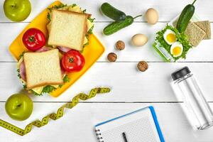 Haut vue Planification carnet avec copie espace et en bonne santé petit déjeuner. sandwich avec jambon et fromage, œufs, concombre, tomate et bouteille de l'eau sur blanc en bois tableau. photo