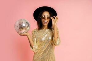 célébrer femme dans d'or paillettes robe posant avec disco ballon plus de rose bacground dans studio. photo