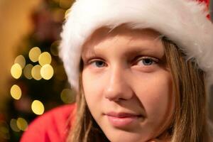 portrait de adolescent fille dans Père Noël chapeau séance sur chaise dans de face de Noël arbre, joyeux Noël et content vacances photo