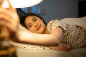 asiatique femme mensonge sur le lit tournant de le lumière à le chevet à sommeil ce soir. photo