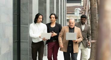 groupe de affaires gens en marchant et travail sur portable ordinateur dans côté de moderne Bureau bâtiment. Multi-éthnique groupe de affaires gens en utilisant numérique dispositifs Extérieur. photo