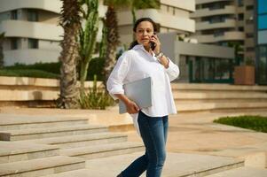 Jeune adulte femme parlant sur mobile téléphone et à la recherche dans le distance, en marchant vers le bas le pas contre paysage urbain photo