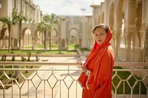 charmant musulman femme dans Orange foulard, à la recherche à caméra, permanent sur le balcon contre le mosquée jardin Contexte photo