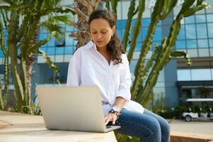 Latin américain Jeune femme étudiant dans bleu jeans et blanc chemise, dactylographie texte sur ordinateur portable, en ligne travaux sur diplôme projet photo