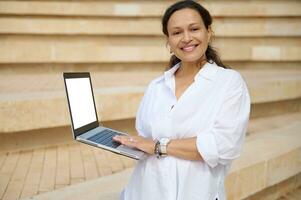 agréable souriant Jeune femme à la recherche à caméra, en portant portable avec blanc maquette numérique filtrer. copie La publicité espace photo