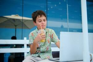 sur de soi intelligent élémentaire âge étudiant garçon en buvant jus et Faire devoirs sur ordinateur portable, séance sur une café été terrasse photo