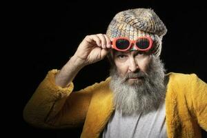 un personnes âgées homme avec une gris barbe, qui est portant une tricoté chapeau et une brillant Jaune fourrure manteau. à la mode homme sur une noir Contexte. photo