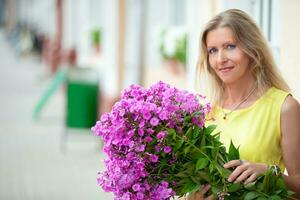 magnifique personnes âgées femme avec une bouquet de fleurs. âge modèle blond avec bleu yeux sourit et regards à le caméra. une content femme dans sa la cinquantaine. photo