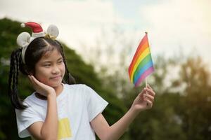 asiatique mignonne fille en portant arc en ciel drapeau, lgbt symbole, flou parc arrière-plan, concept pour lgbt gens fête dans fierté mois autour le monde. photo