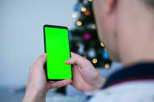 fermer coup de homme main en utilisant mobile téléphone avec vert écran contre le toile de fond de une Noël arbre. doigt balayer en haut et vers le bas dans vert écran de le mobile. vert écran maquette modèle . photo