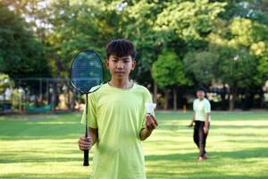 un asiatique garçon détient une badminton raquette et une blanc volant tandis que en jouant badminton avec copains sur le parc pelouse dans le soir après de retour de école. photo