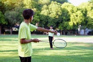 un asiatique garçon détient une badminton raquette et une blanc volant tandis que en jouant badminton avec copains sur le parc pelouse dans le soir après de retour de école. photo