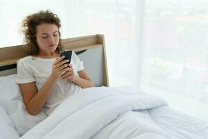 Jeune magnifique content femme relaxant et en utilisant téléphone intelligent tandis que mensonge dans lit dans le Matin après veille en haut. photo