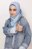 mode portrait de élégant musulman fille dans jean veste donnant les pouces en haut. moderne et Jeune moyen-orient fille dans hijab avec denim veste et Faire content les pouces en haut geste. recommander un service photo